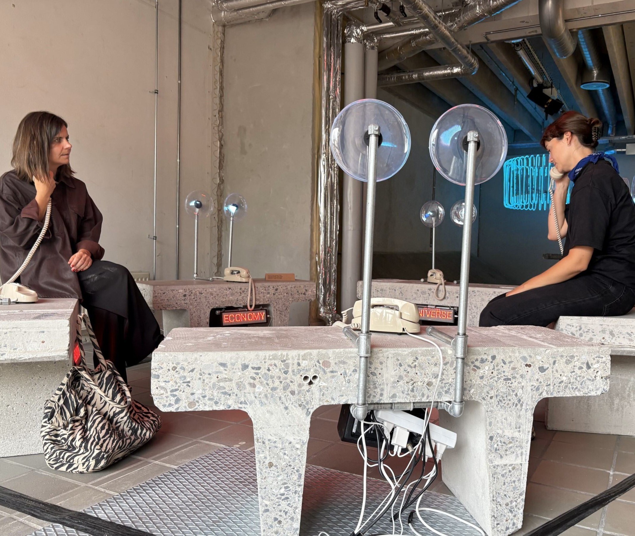 To personer sidder på bænke og interagerer med et kunstværk, hvor man lytter i telefonrør